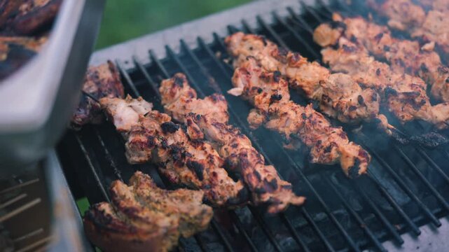 Grilled skewers of marinated chicken cooking on a barbecue grill, smoke rising, with a metal tray holding additional skewers visible in the background