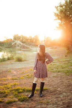 Woman stands on the ground next to a river. The sun sets behind trees, creating a warm light. The person wears a dress and boots