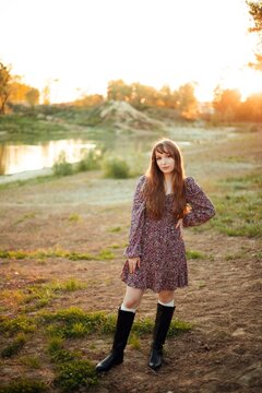 Woman stands on the ground next to a river. The sun sets behind trees, creating a warm light. The person wears a dress and boots