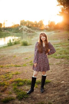 Woman stands on the ground next to a river. The sun sets behind trees, creating a warm light. The person wears a dress and boots