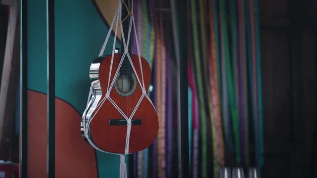 Acoustic guitar suspended with white rope against a colorful backdrop of vertical ribbons and a vibrant mural in a rustic indoor setting
