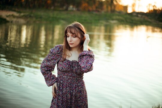 Woman standing by the water during sunset with green trees in the background