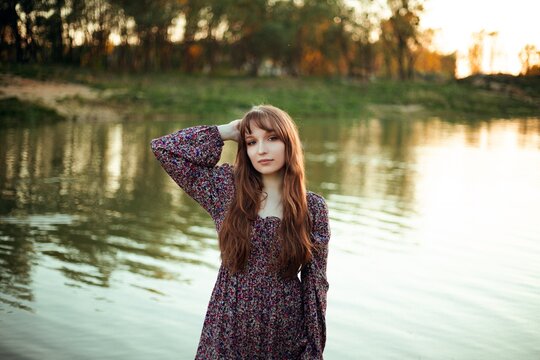 Woman standing by the water during sunset with green trees in the background