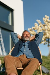 Fototapeta premium Bearded mature man resting outdoors on a sunny slope by contemporary house for relaxation and wellness promotion background