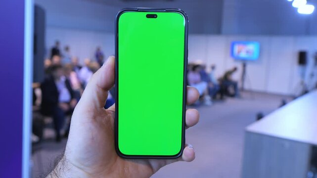 Hand holds smartphone with green screen. Blurred conference room in background. Three keyframes show same scene