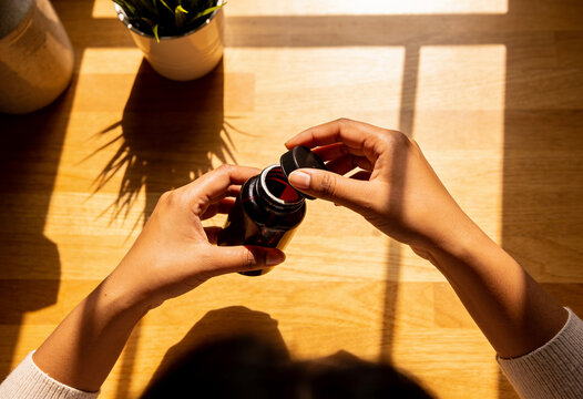 Person hands opening brown medicine bottle on wooden desk warm natural sunlight shadows daily wellness routine health