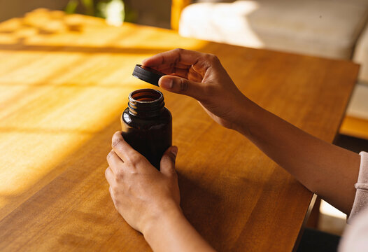 Person hands opening amber glass medicine bottle on wooden table in warm natural sunlight for wellness routine