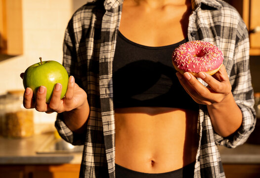 Fitness woman choosing between green Generic Object and pink donut in sunlit kitchen healthy lifestyle nutrition balance