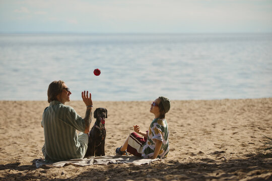 Caucasian father and kid throwing ball to pet dog while relaxing on sandy lake beach. Concept of outdoor family leisure, suitable for summer travel or pet care campaigns
