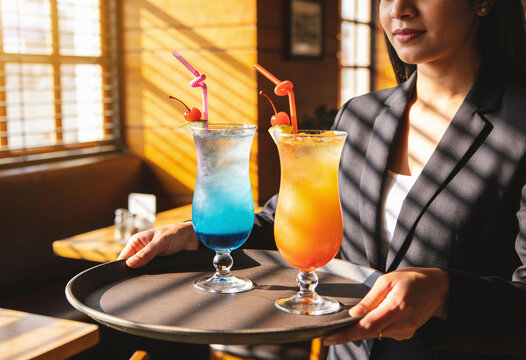 Female server holding tray with blue and orange cocktails in sunlit restaurant with warm natural lighting
