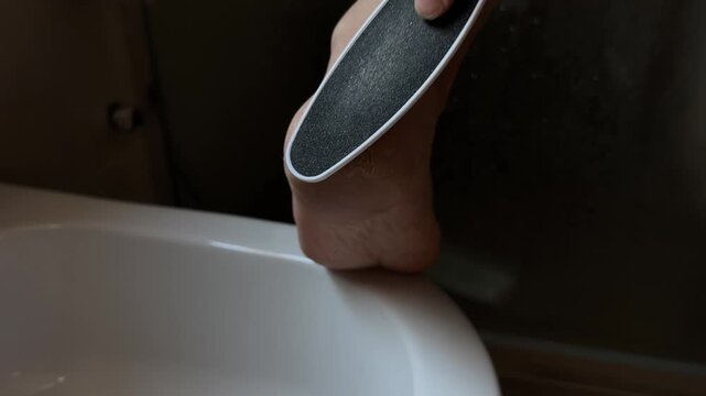 Close up shot of a person using a foot rasp grater to file dry cracked skin on the heel in a dim bathroom setting, focusing on foot care routine and dermatology health.