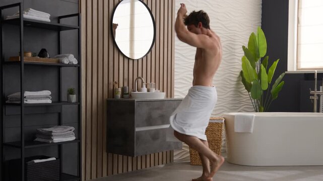 Happy playful Caucasian man dancing in bathroom with towel on hips looking at mirror joyful male fooling around doing morning routine silly funny energetic guy smile having fun singing music indoors