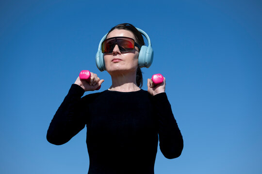Woman wearing headphones and sunglasses against blue sky, working out with hand weights
