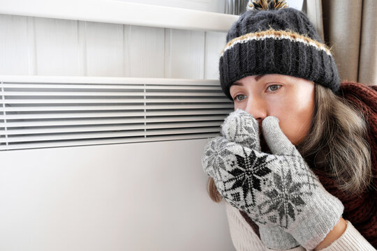 Young frozen woman wearing a sweater shaking and freezing for winter cold with shock expression on face. Girl is warming up hands with breath over electric heater. Discomfort spending time at home