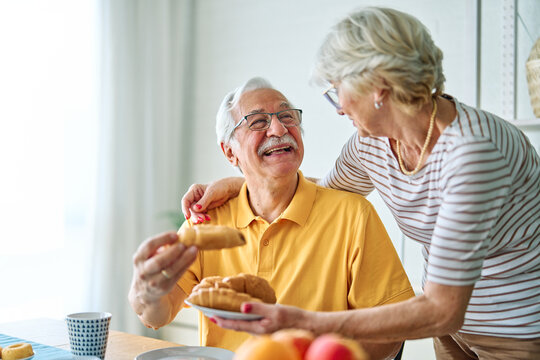 Portrait of an elderly senior couple having breakfast sharing a donut dessert at home. Happy healthy affectionate senior couple eating and sitting at kitchen table having fun enjoying morning meal tog