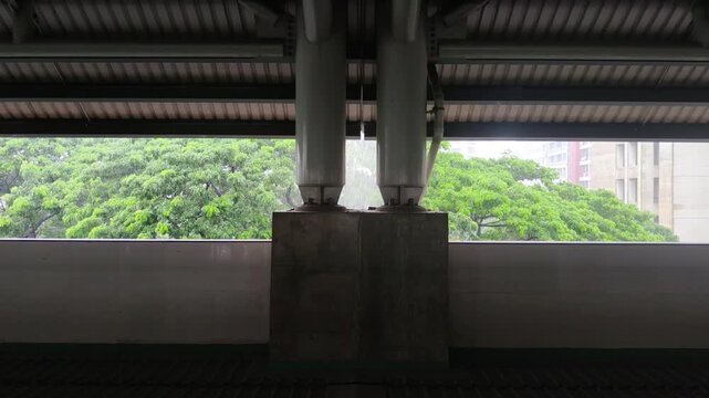 Heavy rain pours outside transit station window with structural steel pillars framing lush green trees in soft daylight showcasing peaceful urban contrast.