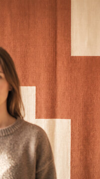 Blurred woman in a knit sweater stands before a woven terracotta and cream textile backdrop