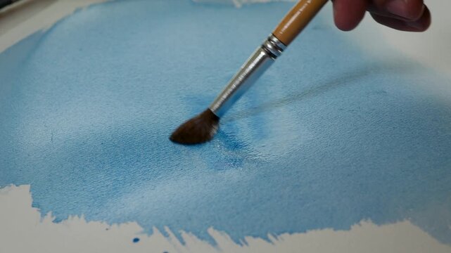 Brush adding blue watercolor to a wet painted surface, creating soft blooms, pigment diffusion and organic marks on textured paper. Creative painting process for artistic and design projects.