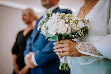 Bride holding elegant white flower bouquet with lace sleeve and ring, accompanied by formally dressed people in softly blurred indoor setting, wedding ceremony portrait © Raivo