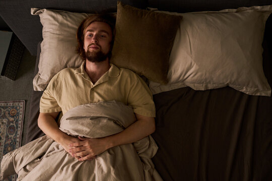 High angle shot of Caucasian young adult man lying awake in bed under blanket. Image suitable for sleep disorder awareness, insomnia treatment campaigns, mental health advertising