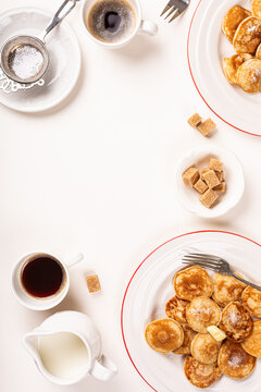 Dutch poffertjes topped with butter sit on red rimmed plates beside coffee, tea, milk, sugar cubes, and utensils, creating a bright overhead breakfast arrangement on a white surface with copy space
