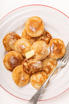 Dutch poffertjes mini pancakes are arranged on a white plate with red trim, dusted with sugar and topped with butter, while a fork rests along the right side in an overhead view