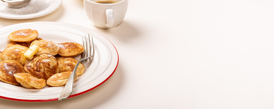 Small golden Dutch poffertjes pancakes topped with butter sit on a white plate with a red rim, while a fork and coffee cup rest nearby. Banner with copy space