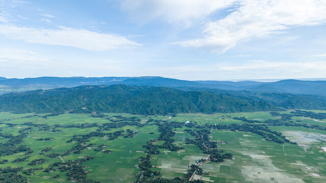 Aerial view of lush green agricultural fields and small villages nestled at the foot of a majestic mountain range under a clear blue sky
