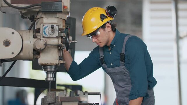 Young man wears hard hat and safety glasses. He operates a milling machine with focus. His hands adjust settings with precision