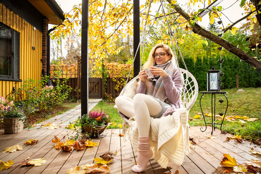 A mid adult woman enjoys a warm drink in a swinging macrame chair on a cozy autumn patio. Mental clarity outdoors during a mindful routine.