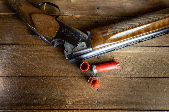 Over Under Shotgun and Shells, An open over under shotgun with polished wooden stock and engraved metal receiver rests on a rustic wooden surface beside two red shell casings