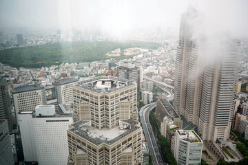 霧に包まれた東京、西新宿の高層ビル群と代々木公園の広大な緑を俯瞰した都市風景。低い雲がビルを覆う幻想的で静かな雨の日の都会の情景。 © BRUCE