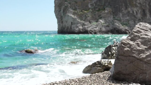 Turquoise ocean beach waves crashing against a rocky shore with a large cliff in the background during a bright sunny day