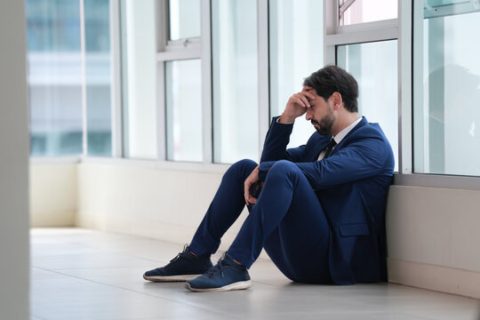 Businessman sitting on office floor beside window, holding head with concern and fatigue during stressful workday in modern workplace