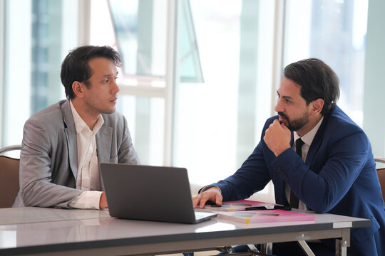 Asian businessman discussing with bearded colleague beside laptop at bright office table, showing focused collaboration during professional workplace meeting