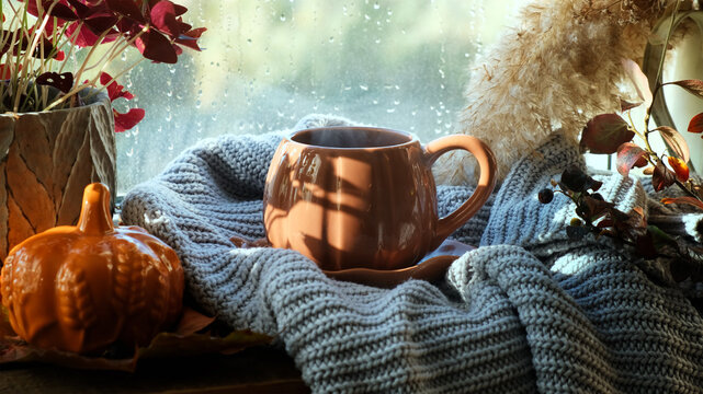 Autumn vibes. Tea time or coffee break. Mug of hot steaming drink on windowsill in cozy interior, raining outside. Warm cozy fall mood. 