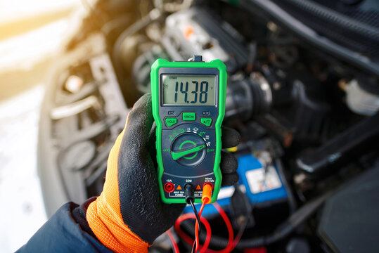 Gloved hand holding digital multimeter showing 14.38 volts while testing car battery under open hood during cold winter weather. Properly functioning alternator, winter electrical system inspection