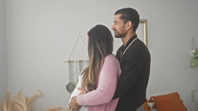 Pregnant woman embraced from behind by man, hands on belly in a cozy home living room with boho wall hanging and plants; tenderness anticipation.