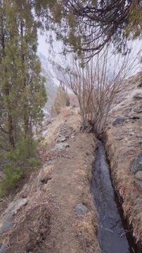 Vertical POV walking along narrow dirt trail and irrigation water canal in arid rocky mountain landscape, steep mountain ridge path next to flowing water stream and bare winter trees