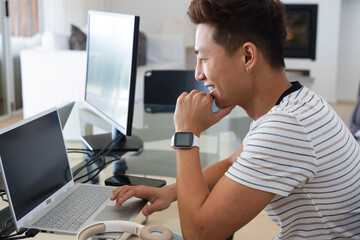 Fototapeta premium Asian man sitting at glass-topped desk working on laptop and monitor, smiling, copy space
