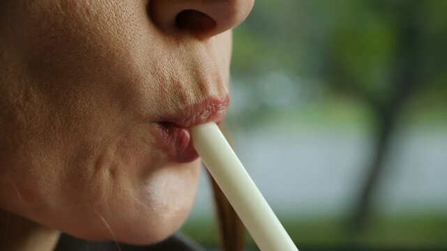 Extreme close up young woman lips drinking cold beverage through white plastic straw, showing suction, enjoyment refreshing milkshake on summer day in cafe. Woman drinking milkshake from plastic straw
