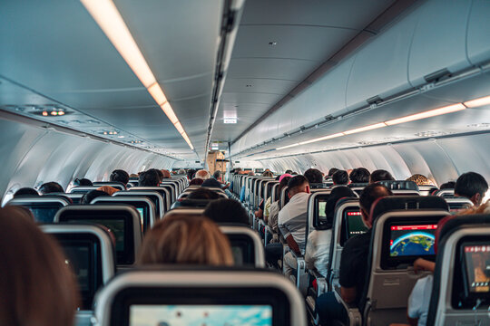 A packed airplane cabin shows passengers seated closely together, each engaged with their personal screens. The image captures the confined yet organized environment of air travel.