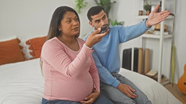 Pregnant woman holds tv remote on bed, hand on belly, partner gestures with open hand beside her; relationship tension.