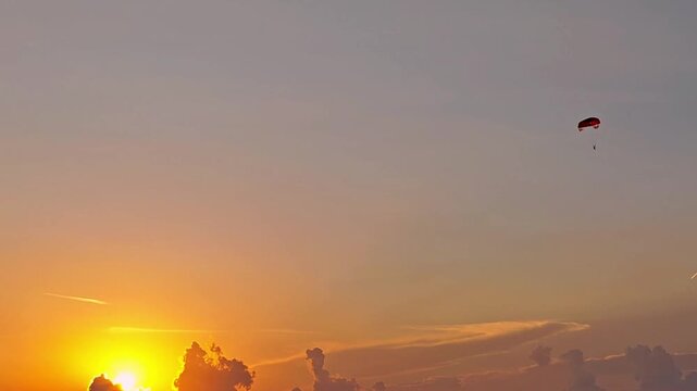 Paraglider descending across glowing sunset sky above clouds with warm orange light and distant atmospheric horizon during tranquil evening aerial flight
