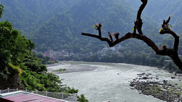 Pruned branches frame a bend of the Ganga River and wooded mountains above a red roof in Rishikesh.