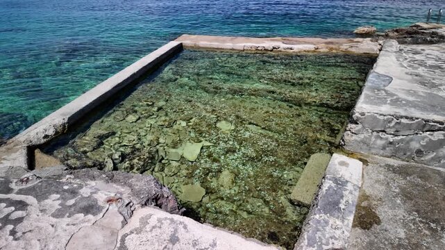 Clear turquoise water washing over natural rock pools and concrete seawalls along the waterfront harbor in Luka Rovenska, Losinj Island, Croatia, offering a refreshing summer swimming experience