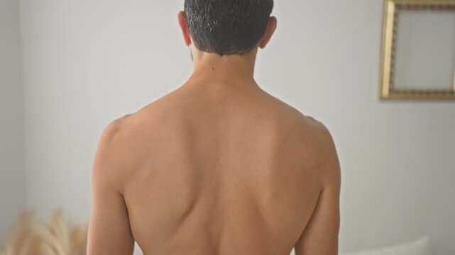 Man shirtless showing bare back and spine standing facing away in a studio bedroom with pillow, bed, pampas grass and empty frame visible; calm introspection.
