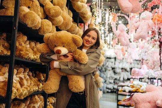 Woman holding oversized teddy bear in a colorful plush toy store aisle surrounded by shelves of stuffed animals and hanging pastel decorations indoors