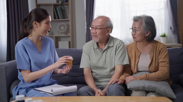 Asian nurse explains medication to an elderly couple sitting together on a sofa in a living room, providing home healthcare and medical consultation with care, empathy, and professional support.
