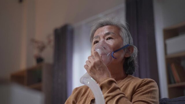 Asian elderly woman uses a nebulizer mask to inhale medication at home, focusing on respiratory health, coping with chronic illness, and managing breathing difficulties with medical equipment.